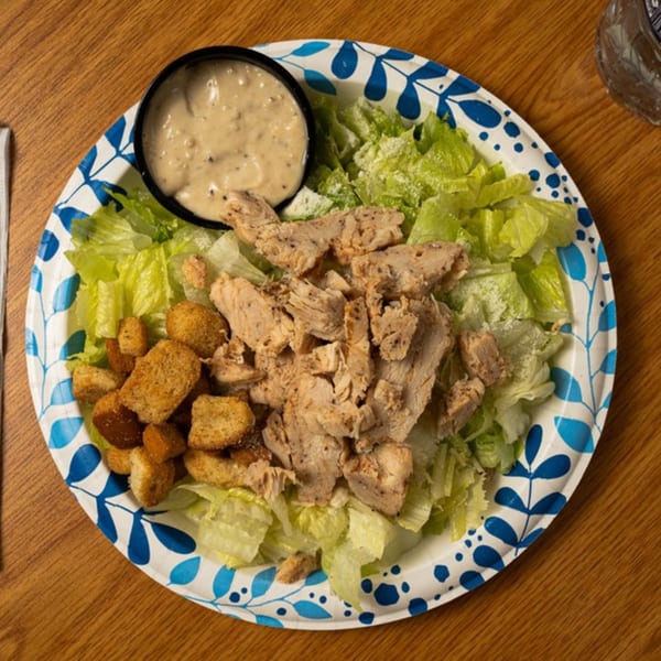 Chicken Caesar salad with croutons and dressing in a cup on a blue and white paper plate.