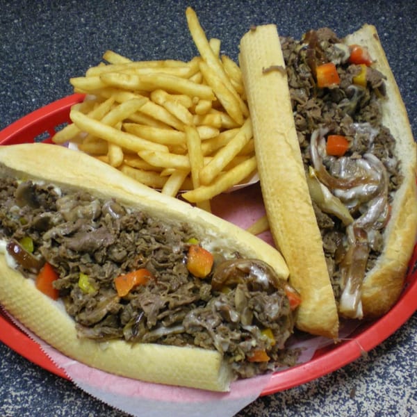 Two Philly cheesesteak sandwiches with fries in a red basket from Phillys Cafe & Deli.