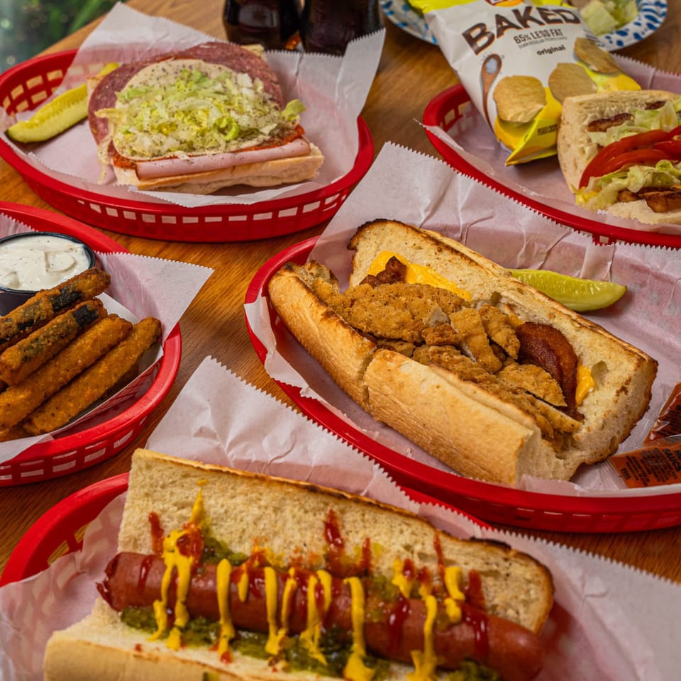 A table laden with various sandwiches, hot dogs, fried pickles, and chips in red baskets.